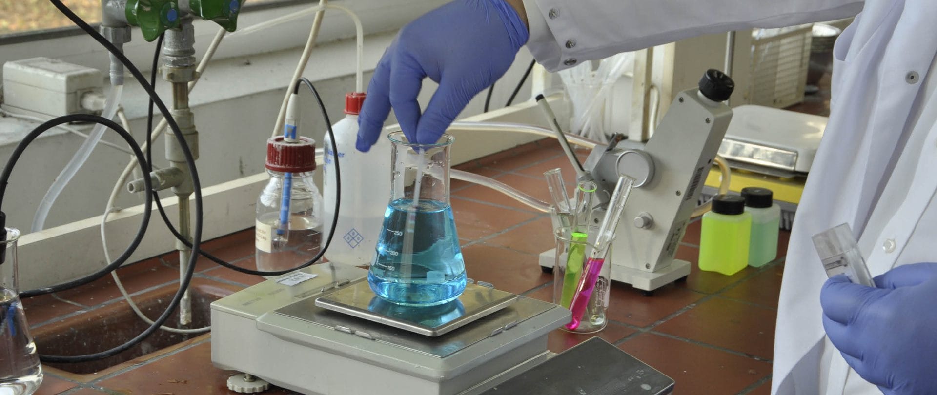 Gloved hand holding a glass stirrer in a conical flask filled with blue liquid on an electronic balance